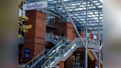 An exterior picture of the Harlequin Theatre in Redhill. There are two sets of stairs up to the theatre, one from the right and the other from the left side. The glass roof has grey panels running both length and width way across.