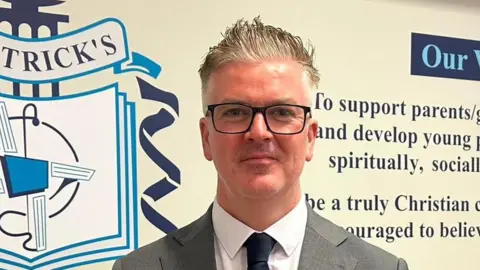 School handout Shows a man in grey jacket with white shirt and black tie standing in front of his school logo