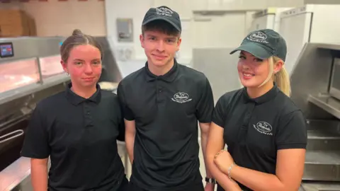 Two young women and one young man (centre) in their chip shop uniforms of a black polo shirt and trousers and a skip cap smile for the camera. They are standing beside the fat fryers and food prep area, all in stainless steel.