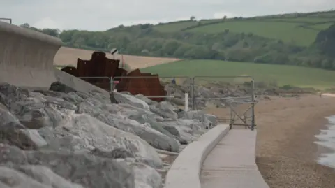 Large rocks and sea defences at Torcross beach. Metal railings are up near where sea defences were damaged. Fields are in the background.