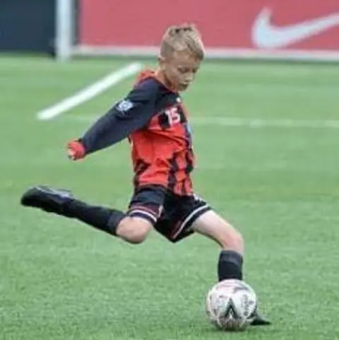 Family handout Boy with red and black football kit and short blonde hair kicking a football on a football pitch.