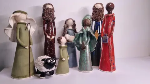 Nativity models made of clay. On the left is a Shepard, wearing green clothes and holding a Shepards crook. To the right of the Shepard is a small sheep. To the right is model of Joseph, wearing brown clothes and with brown hair and a brown beard. To the right is a smaller model of a girl wearing a green dress and with brown hair. To the right is a model of Mary, with brown hair, and holding a baby. To the right are three wise men, one in blue and brown, one in blue and one in red. They are all holding gifts. None of the models have faces. 