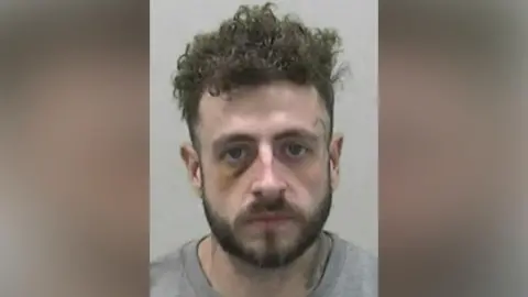 Mugshot of Coe. He has a short dark beard and short thick curly dark hair. He appears to have a bruise around his right eye and an illegible black symbol tattooed beside his left eye