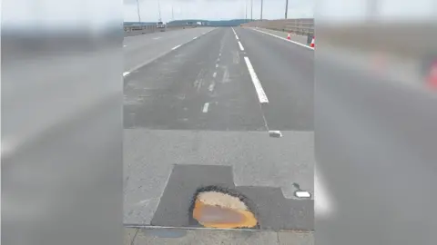 National Highways The M5 motorway on the Avonmouth Bridge. At the bottom of the picture there is a large pothole that has formed on the tarmac, with darker patches of repair work previously done around it. 