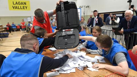A man empties voting slips from a ballot box onto a table with four people wearing blue bibs sitting at it. Photo taken inside the Lagan Valley constituency count centre at South Lake Leisure Centre in Craigavon. 