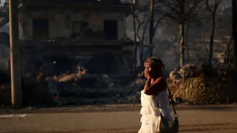 Reuters A woman walks near a burnt house during a wildfire on Salamina island, near Athens, Greece