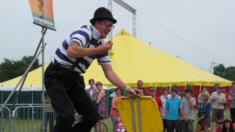 Coventry City Council A clown at the festival in 2019. He is leaning forwards while his left hand is leaning on a yellow plastic square which appears to be anchored to the ground. He has a blue and white striped top with braces over the top connected to his black trousers, a black domed hat and glasses and is smiling. Behind him a small crowd can be seen including children and behind them is a large yellow tent