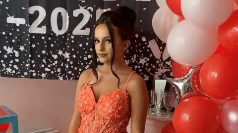 Jessica Metters wearing a pink prom dess. She has long dark hair, tied back, and is standing next to a bunch of white and red balloons.