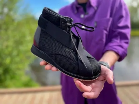 A close-up image of Jeanette holding the show - it is a boot-like style in a breathable mesh fabric, with a rubber sole and an elastic velcro strip starting at the toe, crossing over at the bottom of the laces and stretching around the heel