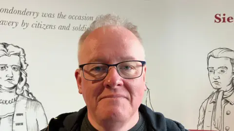 Jonathan Burgess is in a black hoodie and wearing glasses. 
He has short hair and is standing behind a white board with information and historical figures on it. 