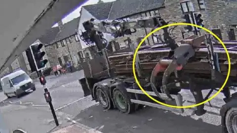 Cambridgeshire Police A CCTV image of a lorry with a large grabber crane on it driving down a road. There is a set of traffic lights and a white van on the left of the image. A yellow circle has been drawn around the crane's grabber.