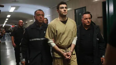 Mangione walking, in jail outfit and handcuffs, led by two guards