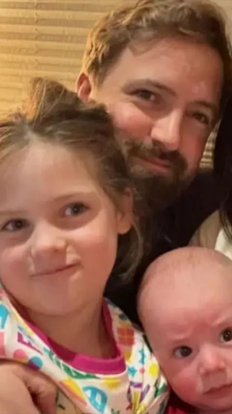Man with a beard smiling and holding a girl in a colourful top and baby boy