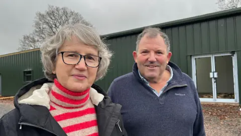 Alison and Peter standing side by side, both smiling and looking into the camera. Alison, on the left, is wearing a red and brown striped roll-neck jumper, a black hooded winter coat and has silver hair cut into a bob. She's also wearing dark framed glasses. Peter, on the right, is wearing a light blue collared shirt with a dark blue fleece over the top. They are both standing in front of a large, single-storey dark green building. The sky is grey and overcast. 
