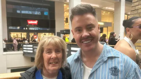 Liguori has short blonde hair and a fringe, she is standing beside her son Luca. He has short dark hair and is wearing a blue shirt with white stripes and a white vest.