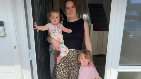 This photograph shows a woman standing in the doorway of a house with two young children. One child is being held in the woman's arms and is wearing a light-coloured outfit. A second child is standing beside the adult, wearing a pink top and light-coloured leggings. The adult is wearing a dark sleeveless top, patterned trousers and dark footwear.