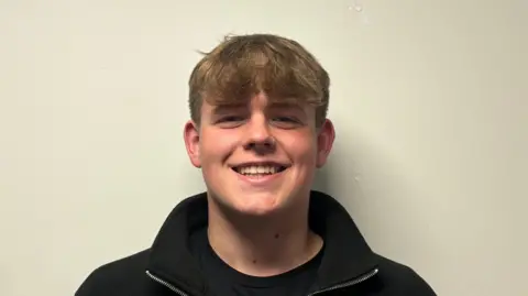 Oscar is standing in front of a white wall. He is wearing a black jumper and black t-shirt. He has brown hair. 