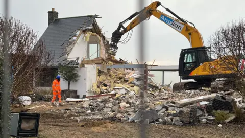 A digger being used to demolish a house hanging on the edge of Thorpeness in Suffolk.