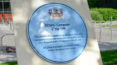 A blue plaque is placed on a white plinth. It has details of the creation of the Edsac computer printed on it.