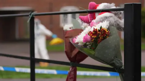 PA Media Flowers and a grey teddy bear tied to a black fence at the suspected murder scene. A police forensic officer, wearing all-white, places a yellow cone on the floor in the background.