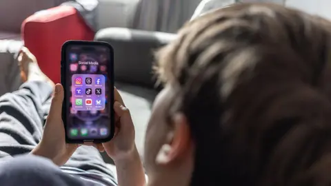 Getty A young boy with mousy-brown hair is lying on a couch and looks at his phone, which says "Social Media" above nine social media apps. 