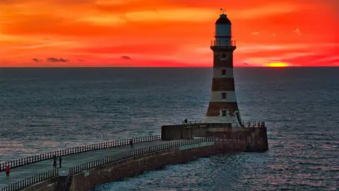 BBC Weather Watchers/Skylark Roker Pier which is a long stone walkway into the sea. The lighthouse stands at the end of it with white and brown-looking stripes and is in shadow. There are people walking across the pier. The sea is calm around it but the sky is a bright red and orange colour.