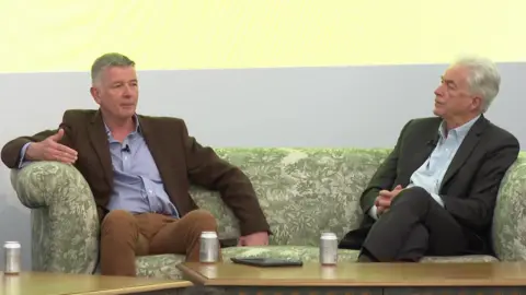 Sir Richard Moore, MI6 chief, wearing a brown suit, sits on a green patterned sofa with William Burns, head of the CIA, who is wearing a dark suit and has has hands clasped.