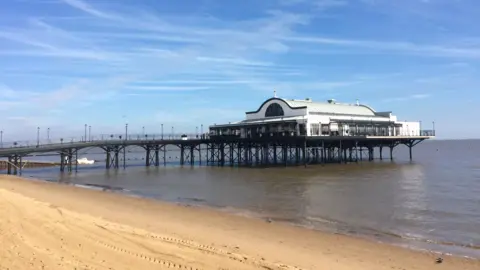 A pier, topped with a large white building with arched roof, juts out into the sea beside a golden beach under a blue sky.