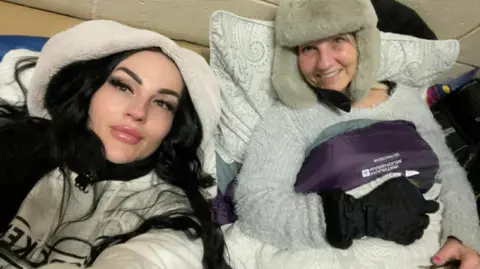 Elaine Barter Elaine wearing a white hoodie, taking a selfie with her mum Linda in a grey jumper and a beige hat. They are lying down. 