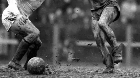 Getty Images Black and white image of two footballers' boots kicking up pieces of mud as they chase the ball