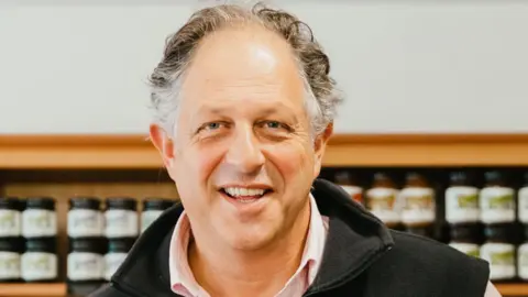 Guy Tullberg  smiles at the camera wearing a black fleece. Behind him slightly blurred is a shelf of condiment jars.