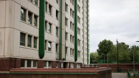 LDRS / Declan Carey A tower block is white and green. A brick wall with a handrail can be seen outside, along with a paved area and trees.