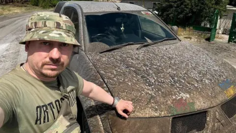 KVPU, the Independent Union of Ukrainian Miners A Ukrainian soldier in a camouflage bucket hat stood in front of a very muddy Mitsubishi SUV. There are Wales flags and Ukraine flags on the front of the vehicle under the dirt