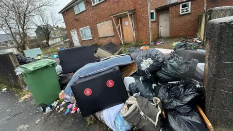 Local Democracy Reporting Service A bed frame and mattress, a sofa and multiple bin bags are piled up in the garden of a house. A green wheelie bin is also placed on the pavement outside the house. The window and front door of the property are boarded up.