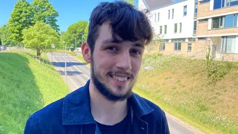 Wilson Colley standing in the sunshine with greenery behind him. He is smiling, with dark hair and a beard and is wearing a dark blue denim jacket