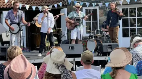 Four men performing as part of a band on stage at an outdoor event. In the crowd are men and women wearing hats.