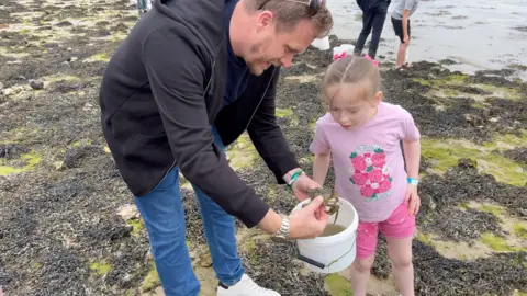 Image shows Lewis with his daughter. They are stood on sand and seaweed, with the water behind them. Lewis is wearing blue jeans and a black zip up hoodie. His daughter is wearing a pink t-shirt and pink shorts. Lewis is holding a white bucket, filled with water and is holding a big crab which they are both looking at. 
