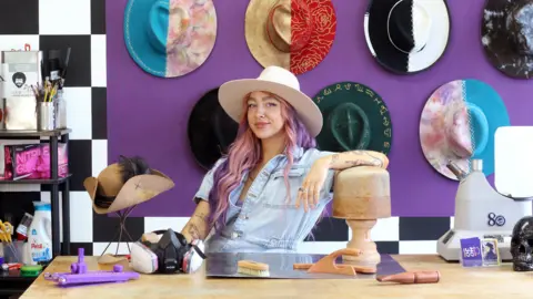 Hojo Hattery/Holly Johnson A woman with long hair which is dyed pink at the front, purple towards the back, looks towards the camera while wearing a cream, wide-brimmed hat. She wears a light blue short-sleeved denim top. She has her left arm resting on top of a circular block of wood on a table. Behind her are a variety of hats hanging on the wall.
