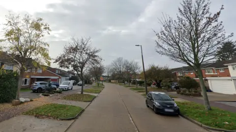 A Google Streetview look at Maplin Way North. A road runs down the middle. Green banks and trees line each side. Then there is a public footpath before the houses which all have large driveways.