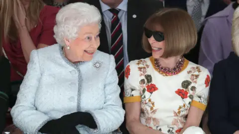 Getty Images Queen Elizabeth II and Anna Wintour sit next to each other and smile at a fashion show in London in 2018