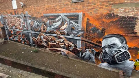James Thompson/Wall Games A street art mural featuring a man with a gas mask and a spray can standing next to some symbols that are painted to look metallic. The background is orange and charcoal.