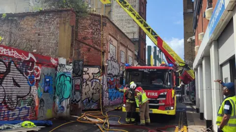 A fire truck and firefighters stand by hoses in a back street in London. There is graffiti on the walls on one side of the road. 