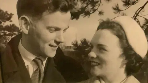 Adrian Hunt A black and white photo of a young man and woman looking at each other affectionately. The man is wearing an overcoat, a light shirt and a striped tie. The woman has short, curly dark hair under a light hat and is wearing lipstick.