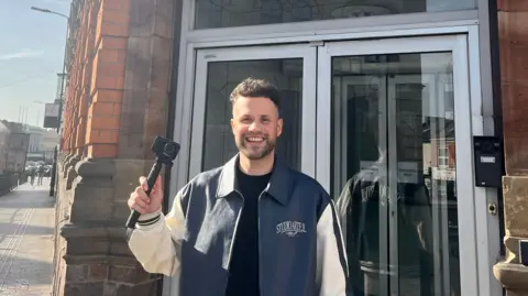 A man stands outside the entrance of BBC Radio Northampton, holding a small camera. They are wearing a dark shirt layered with a blue-and-white varsity-style jacket. The building behind them features red brickwork, large windows. Sunlight casts long shadows across the pavement, and a row of vehicles and buildings lines the street to the left.