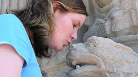 Getty Images A girl leans forward towards the nose of a carved stone gargoyle. The gargoyle has an animal face and fangs and is set against a stone wall with decorative architectural details. The girl has shoulder-length hair and is wearing a light blue top. The stone surface appears weathered, with soft grey tones and worn edges.