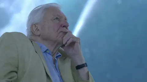 Gavin Thurston David Attenborough is shown in close-up, resting a hand against the chin while looking upward, wearing a light-coloured jacket over a blue shirt. The background is softly lit in cool blue tones.