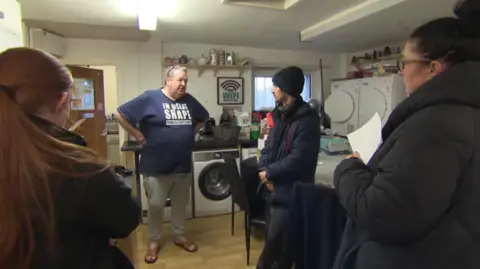 Gerard Woodhouse, wearing a blue t-shirt, sweat pants and sandals, stands in the kitchen of the L6 Centre, where he ran a food bank. He is taking to people who are standing around him. 