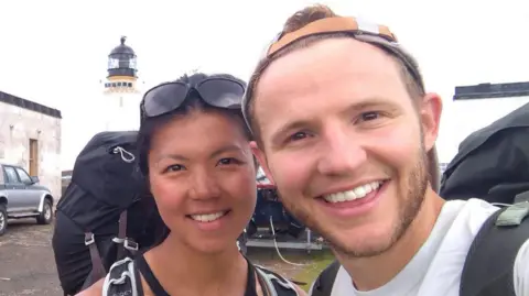 Jade Lau Jade Lau and David Parrish smile at the camera for a selfie. They are standing in a grassy car park with a lighthouse behind them. Jade wears sun glasses and carries a large bag pack. David is wearing a backwards cap and also carries a large bag pack.