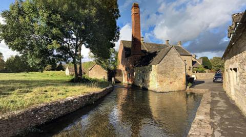 The Old Mill, a quaint brick building with a tall chimney and wooden paddle mill. It is positioned right next to the water on a calm sunny day