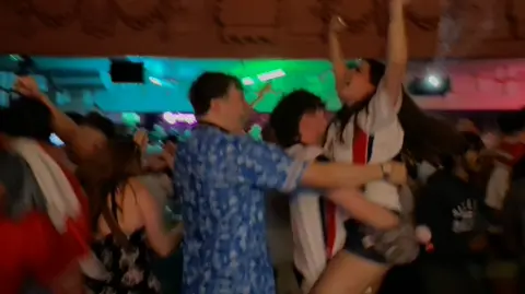 The Grosvenor A woman in a white England jumps into a man's arms as her friend, in a blue England top, also hugs her